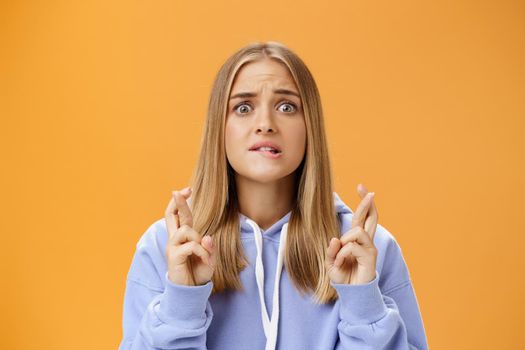 Worried anxious hopeful young woman with fair hair in stylish over-sized hoodie biting lower lip frowning concerned crossing fingers for good luck praying for dream come true against orange wall. Lifestyle.