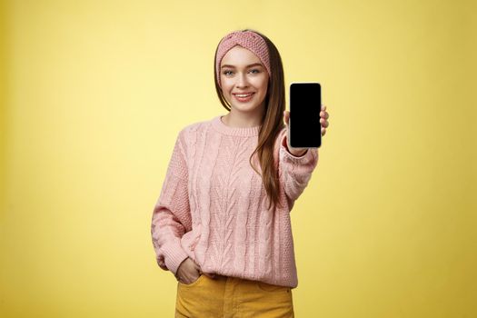 Best smartphone here you go. Charming outgoing young pretty woman in knitted sweater, headband extending hand with phone showing gadget screen smiling recommending cellphone over yellow wall.