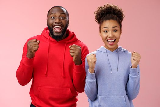 Lucky cheerful two african-american man woman yelling hooray celebrating triumphing huge success clenching fists joyfully accomplish mutual goal standing joyfully pink background victory gesture.