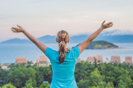 The young woman raised her hand up to the sun, against the sea, rocks and sky with clouds.