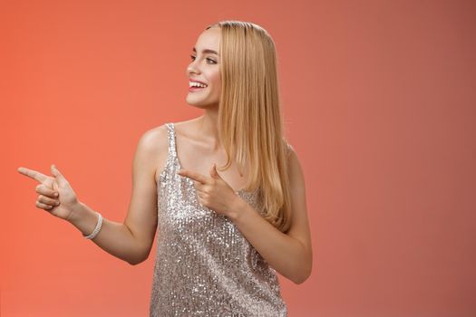 Joyful carefree young wealthy glamour blond woman in glittering silver dress enjoying awesome party dancing turning pointing left smiling broadly waiting girlfriend bring drinks, red background.