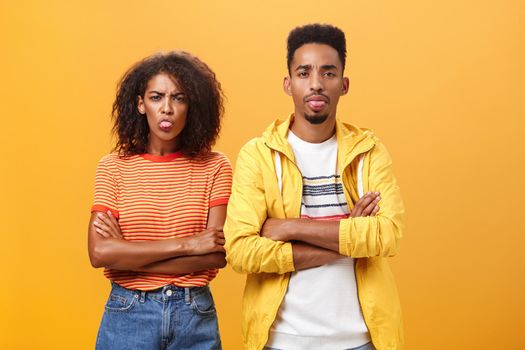 Indoor shot of african american siblings being displeased and annoyed showing bad tempber behaving childish sticking out tongue standing together with crossed hands on chest over orange background. Lifestyle.