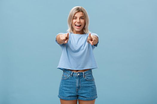 Happy enthusiastic stylish blonde female with tattoos pointing at camera as if picking or making hint smiling joyfully and winking from amazement and happiness posing over blue background. Copy space