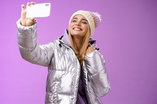Happy delighted carefree fair-haired charming european woman in silver winter jacket hat raising smartphone horizontally taking selfie smiling mobile phone display, purple background.