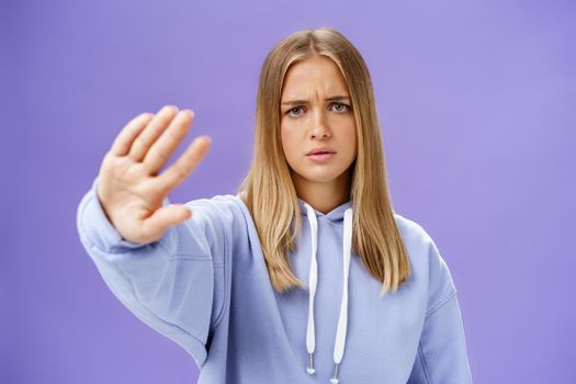 Stop photographing me. Concerned displeased young woman demanding end shooting pulling hand towards camera in no and rejection gesture frowning looking serious and displeased over blue wall. Copy space