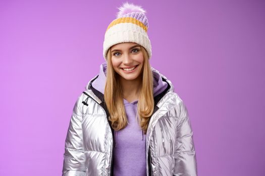 Lifestyle. Cute young blond european woman wearing warm cozy jacket hat ski resort vacation having fun smiling amused rent equipment wanna learn snowboarding standing happily purple background.