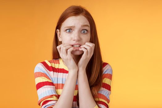 Anxious unconfident timid insecure redhead cute girl worry trembling fear consequences biting fingernails frowning grimacing frightened, standing nervously orange background awaiting scary moment.