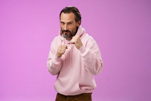 Sassy confident good-looking mature caucasian bearded man in pink hoodie teaching son how stand back fight defend himself raise fists ready boxing smiling daring, stay good shape, purple background.