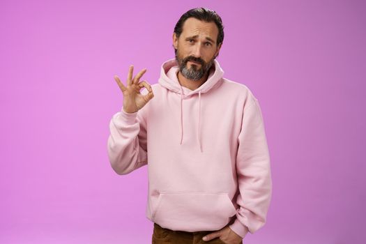Not bad may better. Portrait handsome mature bearded father wearing trendy hoodie frowning awkward show okay ok normal gesture unimpressed but agree, standing purple background doubtful unsure.