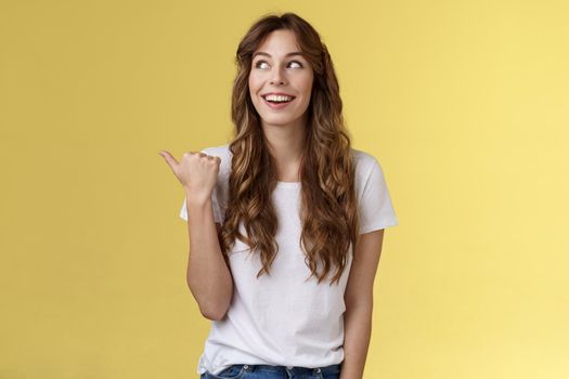Something interesting right there. Curious lively enthusiastic charming woman glancing pointing thumb left smiling broadly discuss cool event laughing carefree stand yellow background.
