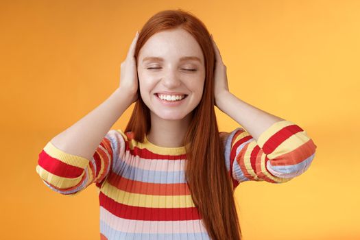 Dreamy charismatic relaxed tender redhead caucasian girl 20s close eyes cover ears imaging far away, smiling broadly enjoy silence peace comfort without sounds noise-free, orange background.