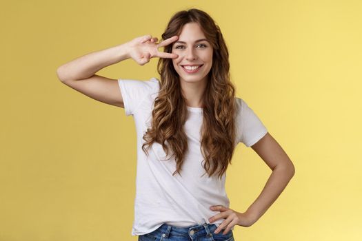 Cheerful positive cute girl curly-haired show peace sign delighted enjoy youth having perfect summer holidays determined rest trip vacation smiling broadly stand confident yellow background. Lifestyle.