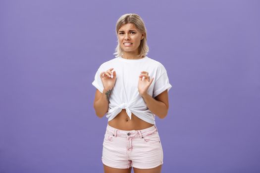 Ew take disgusting thing away from me. Stylish feminine good-looking blond woman in cropped top with tanned skin and pierced belly looking at creepy think with dislike and aversion over purple wall.