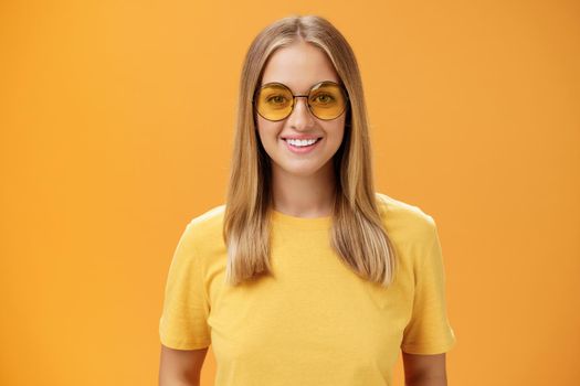 Stylish self-assured charming european woman with fair hair and tanned skin in yellow t-shirt and sunglasses smiling broadly amused and cheerful gazing at camera posing against orange background. Lifestyle.