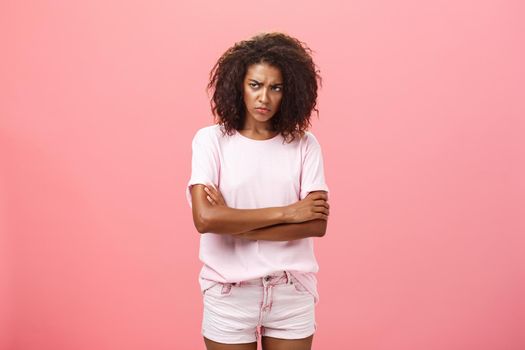 Portrait of offended gloomy sad african american female friend crossing arms on chest in protection gesture frowning looking from under forehead with insult looking envious and angry over pink wall. Lifestyle.