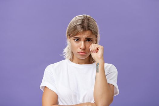 Studio shot of offended sad and timid silly woman with blond hairstyle frowning looking from under forehead holding fist near eye as if whiping teardrop being upset over purple background. Emotions concept