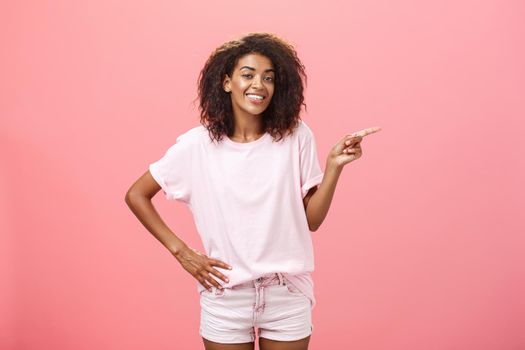 Hey look. Portrait of cute chill and friendly stylish african american woman with afro hairstyle holding hand on waist pointing right and smiling joyfully over pink background. Copy space