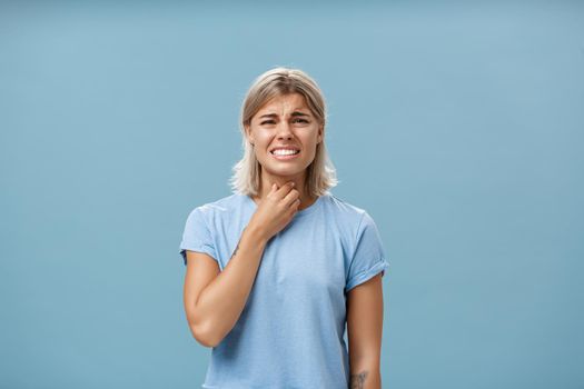 Girl feeling discomfort in throat catching cold or having seasonal allergy touching neck and frowning with clenched teeth and painful expression standing displeased over blue background. Health concept