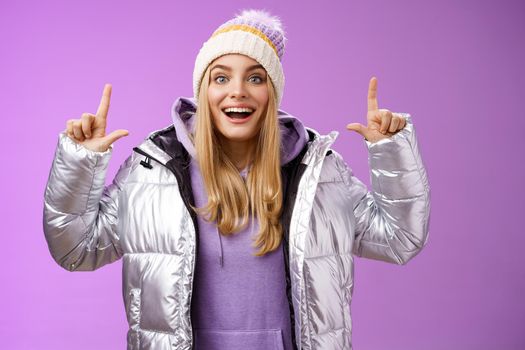 Lifestyle. Excited carefree cheerful fair-haired european girl in silver jacket winter hat raising hands pointing up have excellent idea smiling broadly speaking passionately standing purple background.