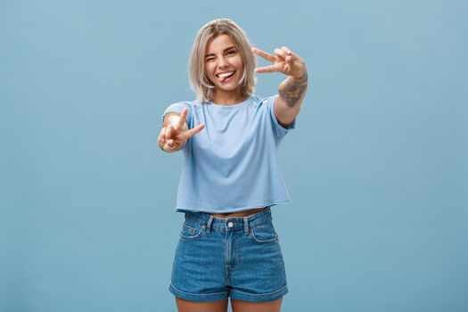 Beautiful tattooed girl enjoying weekends sticking out tongue joyfully winking at camera and smiling showing peace or victory gesture with pulled arms feeling happy posing over blue background. Copy space