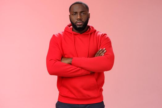 Serious-looking strong black guy security worker cross hands chest confident bossy pose frowning making solid scary look wearing red hooide posing red background, employer controling work.