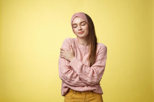 Sweet glamour young girl wearing warm comfy sweater embracing herself crossing arms over body in hug, smiling soft and kind close eyes, recalling lovely days, tender memories over yellow wall.