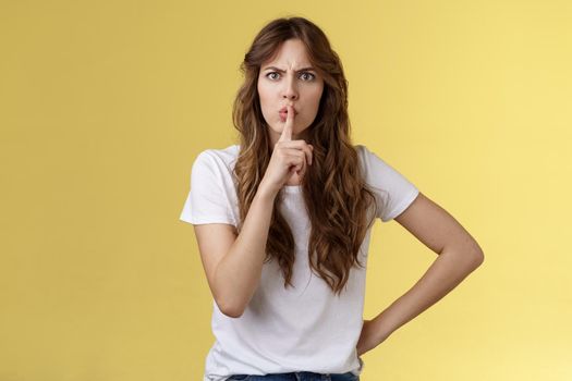 Shut your mouth no talking during concert. Serious-looking strict displeased adult sister demand sit silent hushing hold index finger pressed lips frowning angry shushing yellow background.