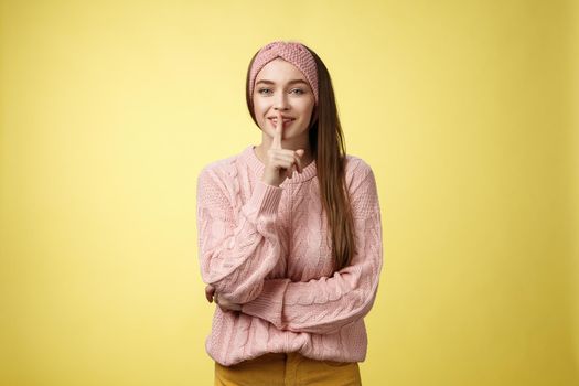 Shh keep voice down. Attractive glamour young stylish schoolgirl having secrets showing shush gesture, holding index finger on mouth smiling, hiding surprise asking not tell anyone over yellow wall.
