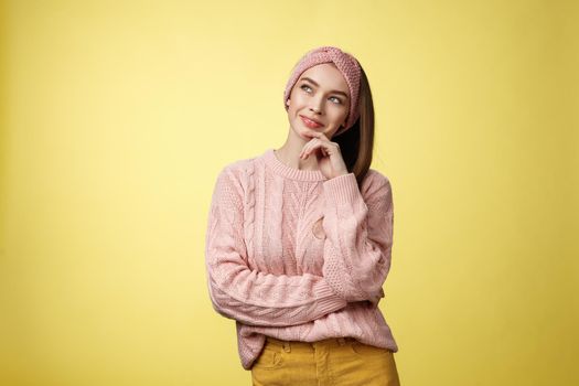 Dreamy attractive and stylish schoolgirl in knitted warm sweater, headband, thinking as smiling satisfied and devious having idea, looking at upper left corner thoughtful, daydreaming, planning.