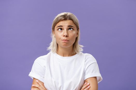 Mind going crazy. Portrait of funny tired attractive young woman with fair hair and tanned skin squinting rolling eyes aside and looking up fooling around over purple background from boredom. Emotions concept