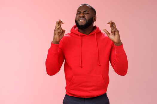 Passionate devoted african-american male fan praying cross fingers good luck asking god win bet biting lip impatiently waiting important results supplicating hopefully, standing pink background.