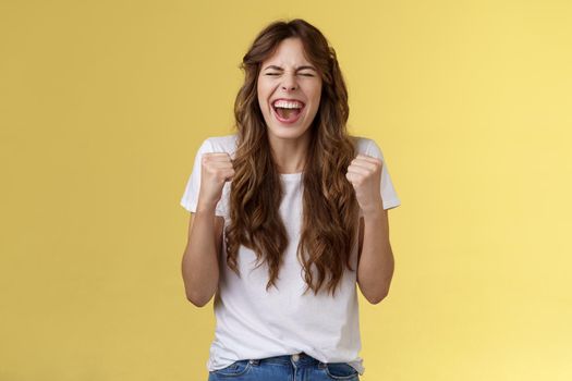 Excited happy triumphing joyful caucasian girl close eyes fist pump celebratory happiness gesture yelling yeah success reach goal achievement dancing victory winning feel relieved yellow background. Lifestyle.