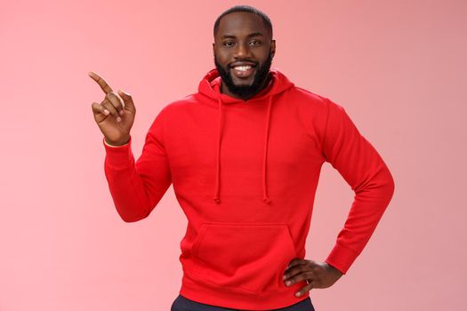 Handsome friendly-looking black guy bearded wearing casual red hoodie pointing left help customer find way directing blank space advertising product smiling happily, pink background.