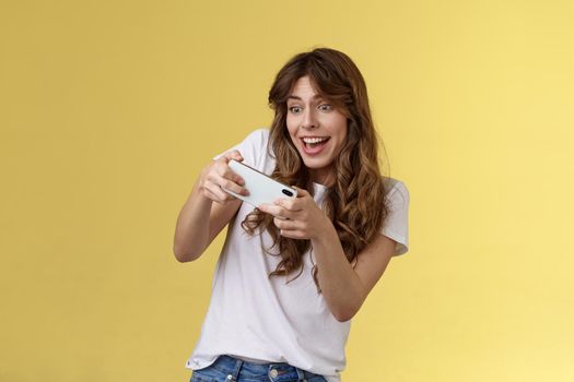 Excited playful enthusiastic girl tilting sideways playing awesome interesting smartphone game car racing smiling determined focuse gaming hold mobile phone horizontal tap display yellow background.