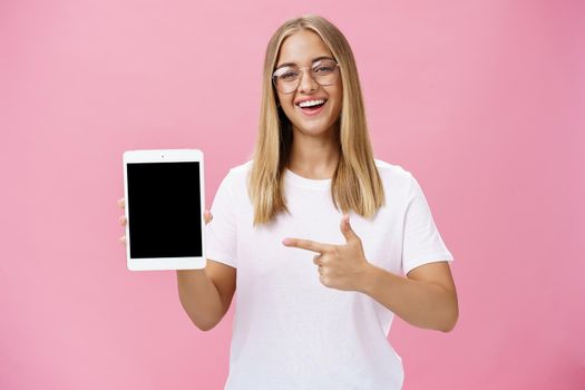 Female freelance programmer proudly showing her app for digital tablet holding gadget pointing at device screen smiling broadly with delighted expression wearing glasses, posing over pink wall. Technology and advertising concept