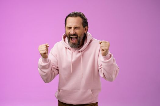 Accomplished happy joyful triumphing mature 40s bearded man grey hair yelling yes thrilled joyfully clench fists celebrating success win lottery become millionaire, standing purple background.