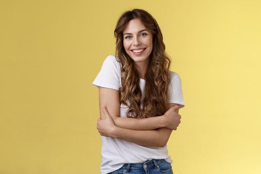 Tender lovely happy good-looking girl long curly hair hug herself wanna warm cuddles smiling toothy pleased friendly conversation having fun wear white t-shirt embracing own body yellow background. Lifestyle.