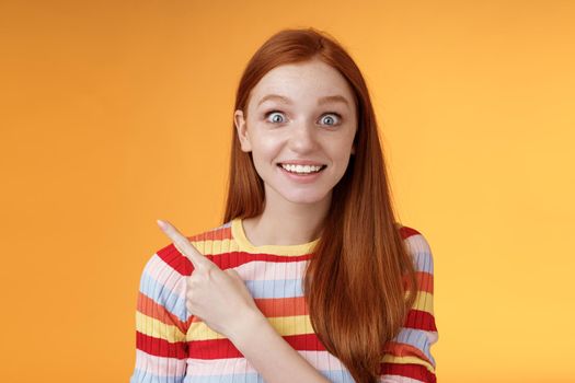 Excited thrilled good-looking young silly redhead girl surprised pop eyes camera gasping impressed pointing upper left corner see super star grinning excitement delight, orange background. Copy space