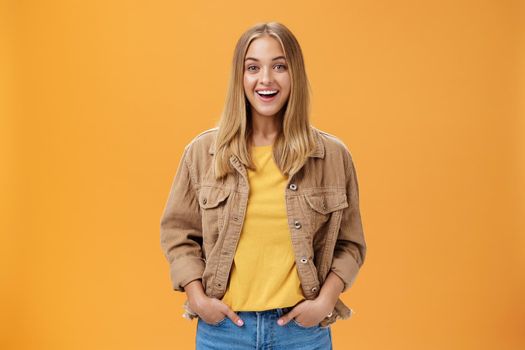 Charismatic tanned woman in corduroy jacket and yellow t-shirt ready for chilly autumn walk with friends smiling joyfully gazing entertained at camera holding hand in pockets casually over orange wall. Lifestyle.
