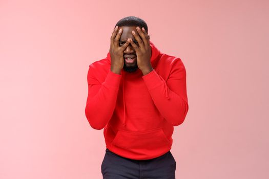 Shocked upset young african american bearded guy in red hoodie facepalm hide face hands peeking through fingers sad devastated lose hope, standing sad pink background grieving feel sorrow.