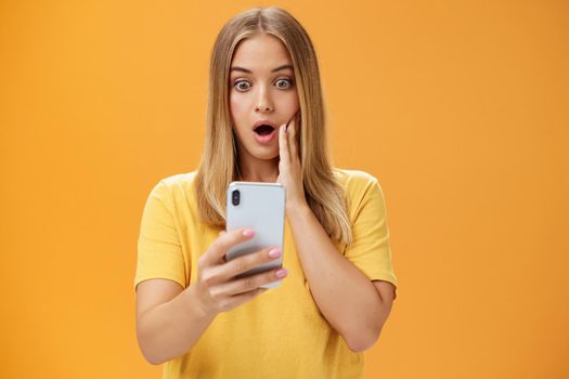 Waist-up shot of shocked and stunned emotive young woman reacting to shocking terrible news reading online from internet gasping opening mouth nervously touchin cheek staring at smartphone screen. Technology concept