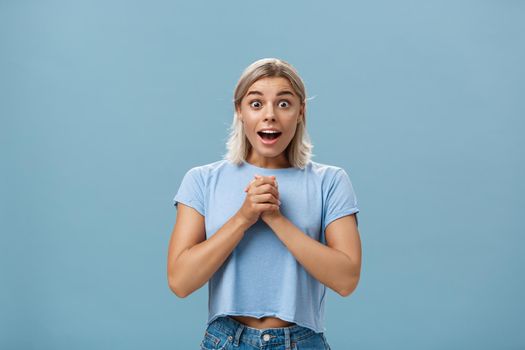 Really is it for me. Portrait of surprised grateful attractive woman with blond haircut clenching hands together on breast opening mouth gasping from admiration and fascination standing over blue wall. Emotions concept