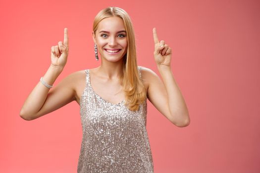 Excited gorgeous blond european woman in silver shiny elegant dress raise hands pointing up showing impressive incredible advertisement smiling happily thrilled wanna take closer look, red background.