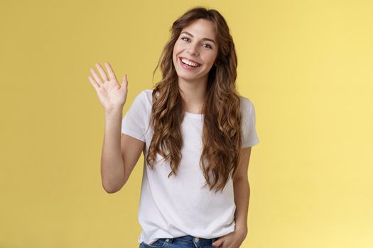 Modest cute friendly outgoing attractive european woman long curly haircut waving palm sociable conversation introduce herself smiling broadly say hi hello greeting coworker yellow background.