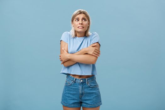 Turn off air-cooler I am freezing in summer. Displeased cute caucasian blonde girl in trendy t-shirt hugging herself while trembling from cold shaking gazing right with worried look over blue wall.