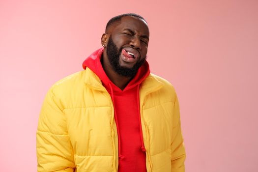 Cheeky stylish funny african bearded man in yellow trendy jacket red hoodie show tongue sassy flirty gaze winking camera trying impress woman pretend macho, standing pink background.