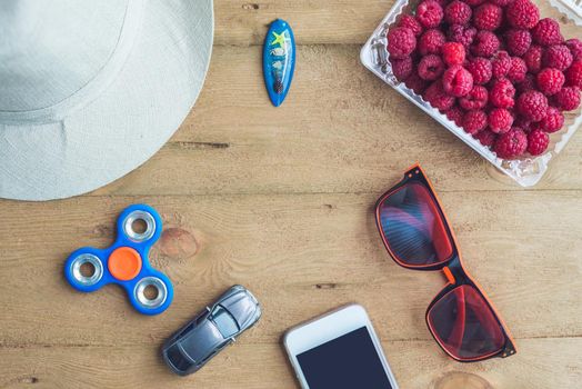 Summer holiday, vacation, relaxation concept. Raspberries, hat, mobile phone, smartphone, spinner, sunglasses from above, top view, flat lay on wooden background. Colorful background with free text copy space. Summer vibes concept.