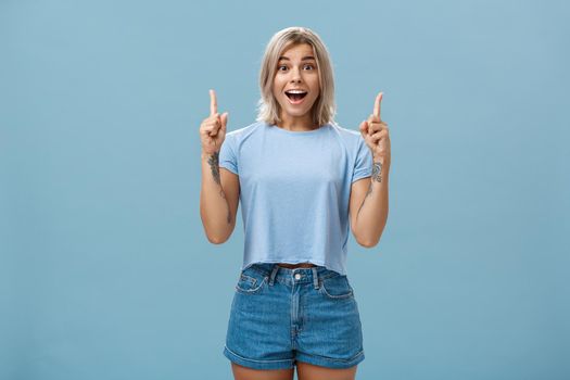 Waist-up shot of impressed enthusiastic creative blonde woman in trendy summer outfit smiling gasping being charmed and thrilled with awesome copy space pointing up over blue background. Copy space