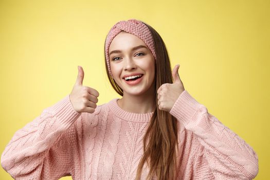 Totally approve. Girl agrees taking part in interesting activity smiling joyful, staying positive showing thumbs up in support, recommend super cool service. posing delighted against yellow wall.