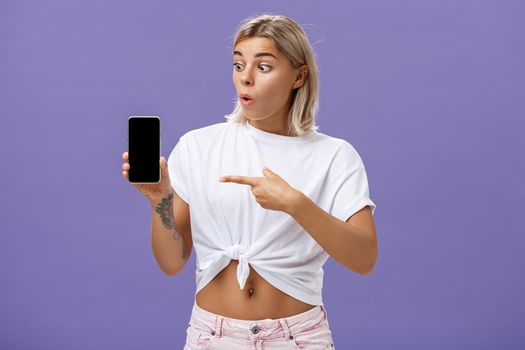 Waist-up shot of impressed and delighted gorgeous european female with blond hair and tattoos looking and pointing at awesome brand new smartphone standing over purple background. Technology concept